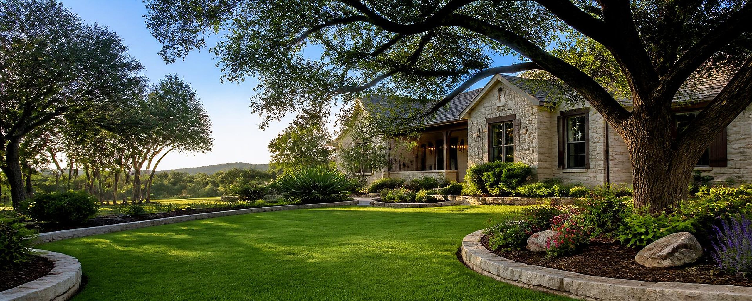 Tidy residential Texas Hill Country landscape with a neat lawn, curved limestone bed borders, mulched planting beds and mature shade trees framing a stone home